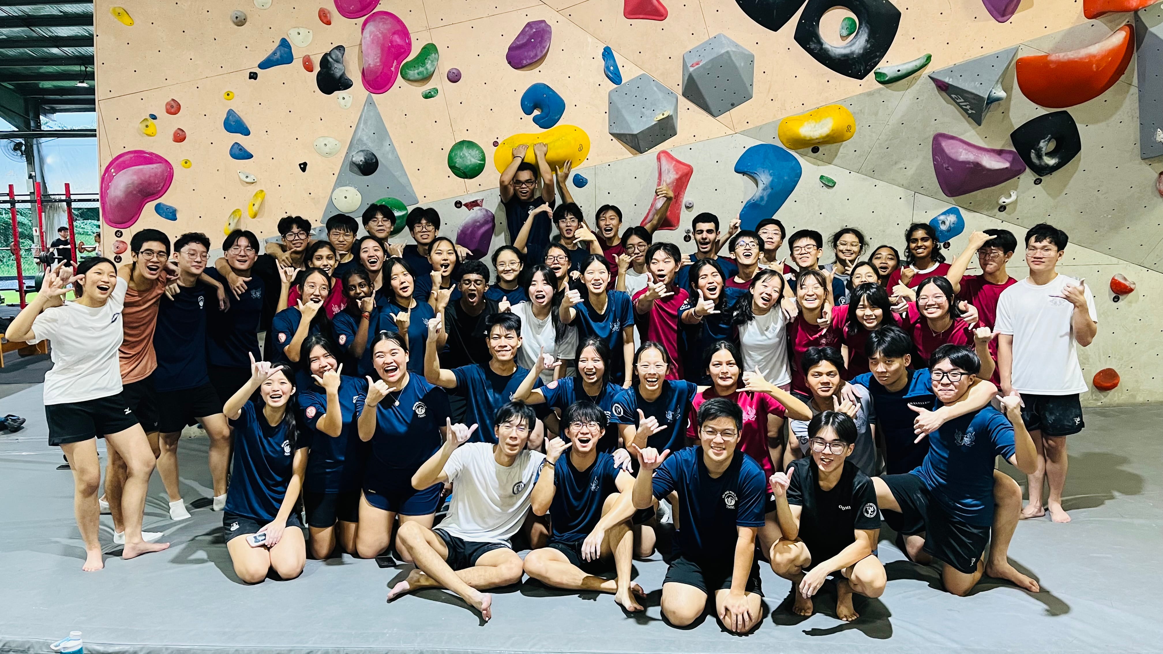 A large group of around 50 smiling and cheering students pose together in front of a colourful indoor bouldering wall.
